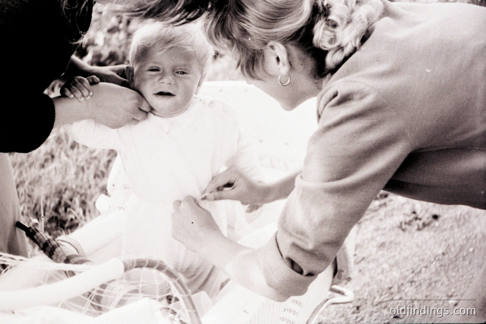 A distressed infant in a white dress is held by a woman with a distinctive hairstyle and hoop earrings. A stroller partially visible in the foreground. Likely a candid moment, potentially 1960s or 70s. Useful for stock photography depicting early childhood or family life.