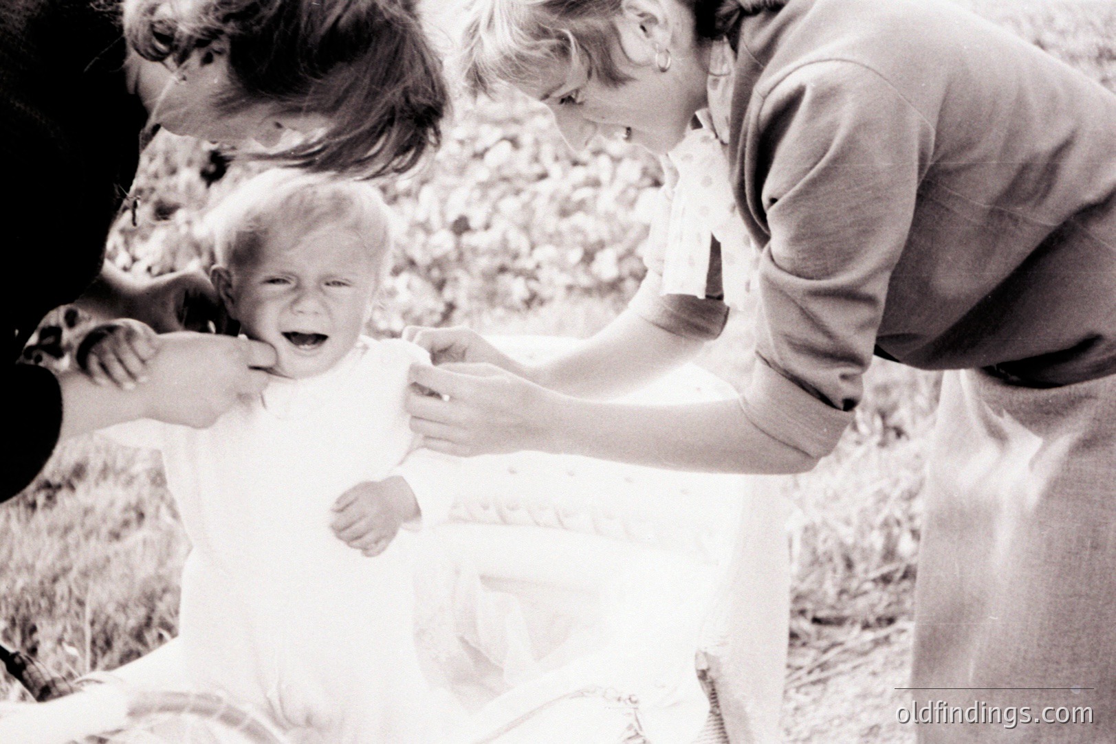 A crying baby in a white dress is being held by two adults. The scene appears to be an outdoor setting, likely a garden or park, with foliage blurred in the background. The adults are dressed in what appears to be 1960s/70s casual attire. Likely a candid snapshot of a family moment.