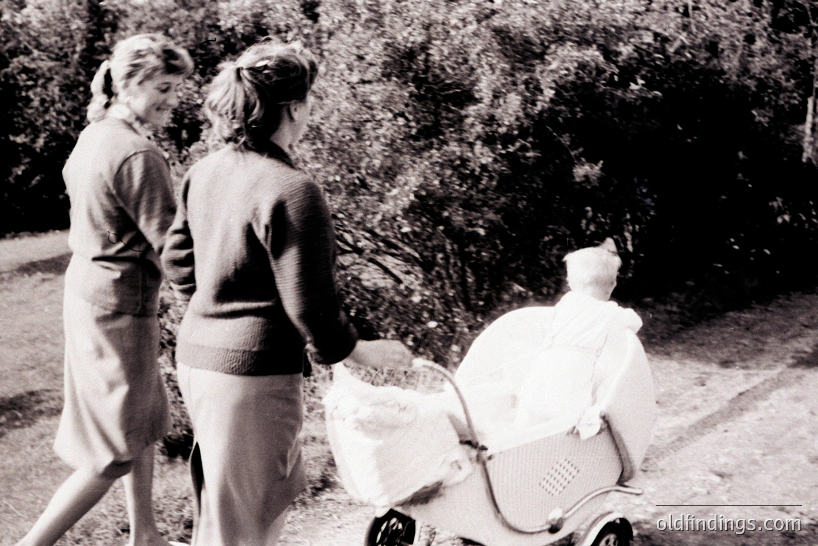 Two women stroll along a path, pushing a vintage pram with a doll inside. The scene appears to be captured in a garden or park setting with foliage visible in the background. Likely mid-20th century, judging by attire and pram style. A charming, nostalgic portrait of childhood.