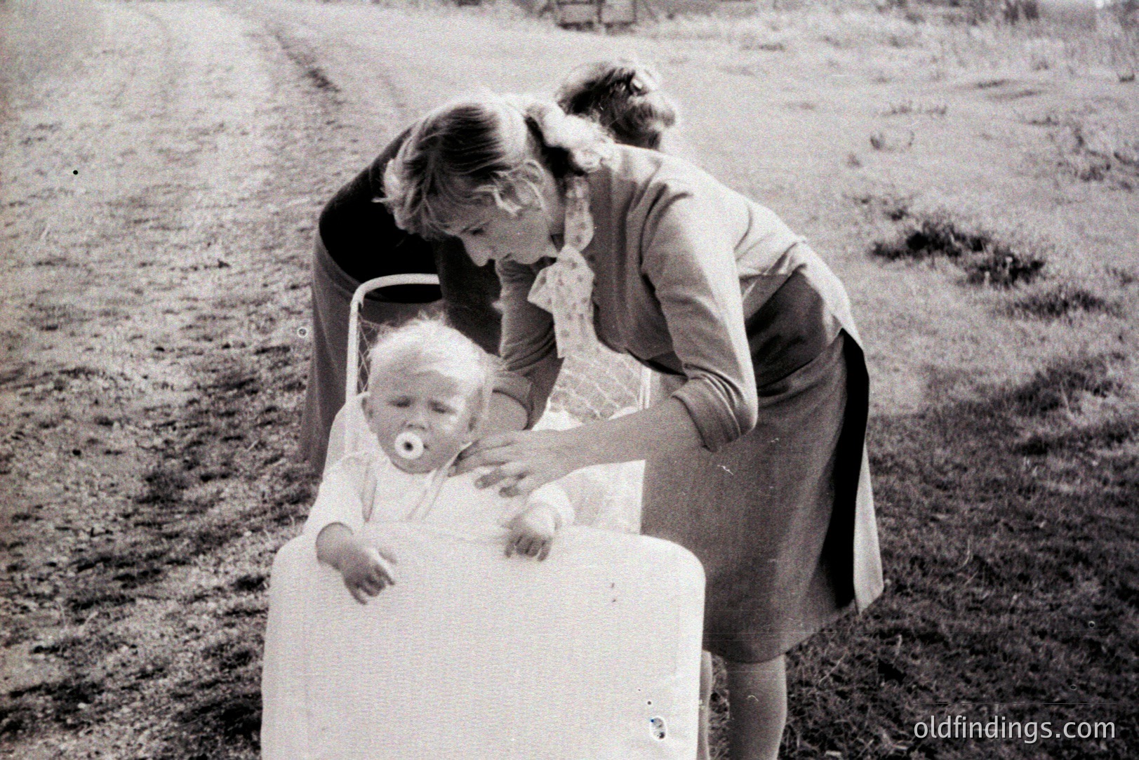 A young child sits in a modified cart or stroller, seemingly outdoors. The caregiver, dressed in a 1960s-style dress and patterned scarf, gestures toward the child. The landscape suggests a rural setting with a dirt track. Likely a candid family moment.