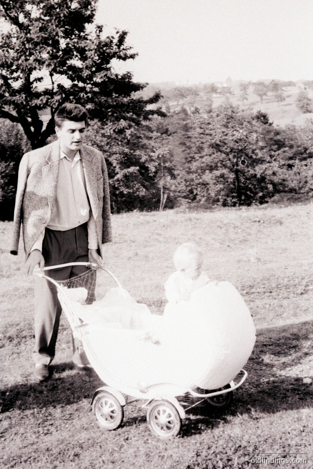 A man in a tweed suit pushes a vintage carriage containing a baby on a grassy field, backed by a gentle hillside. The carriage’s style suggests a mid-20th century aesthetic. Likely captured in the 1950s or 60s, this image captures domestic life. Offers potential for design references and historical research.