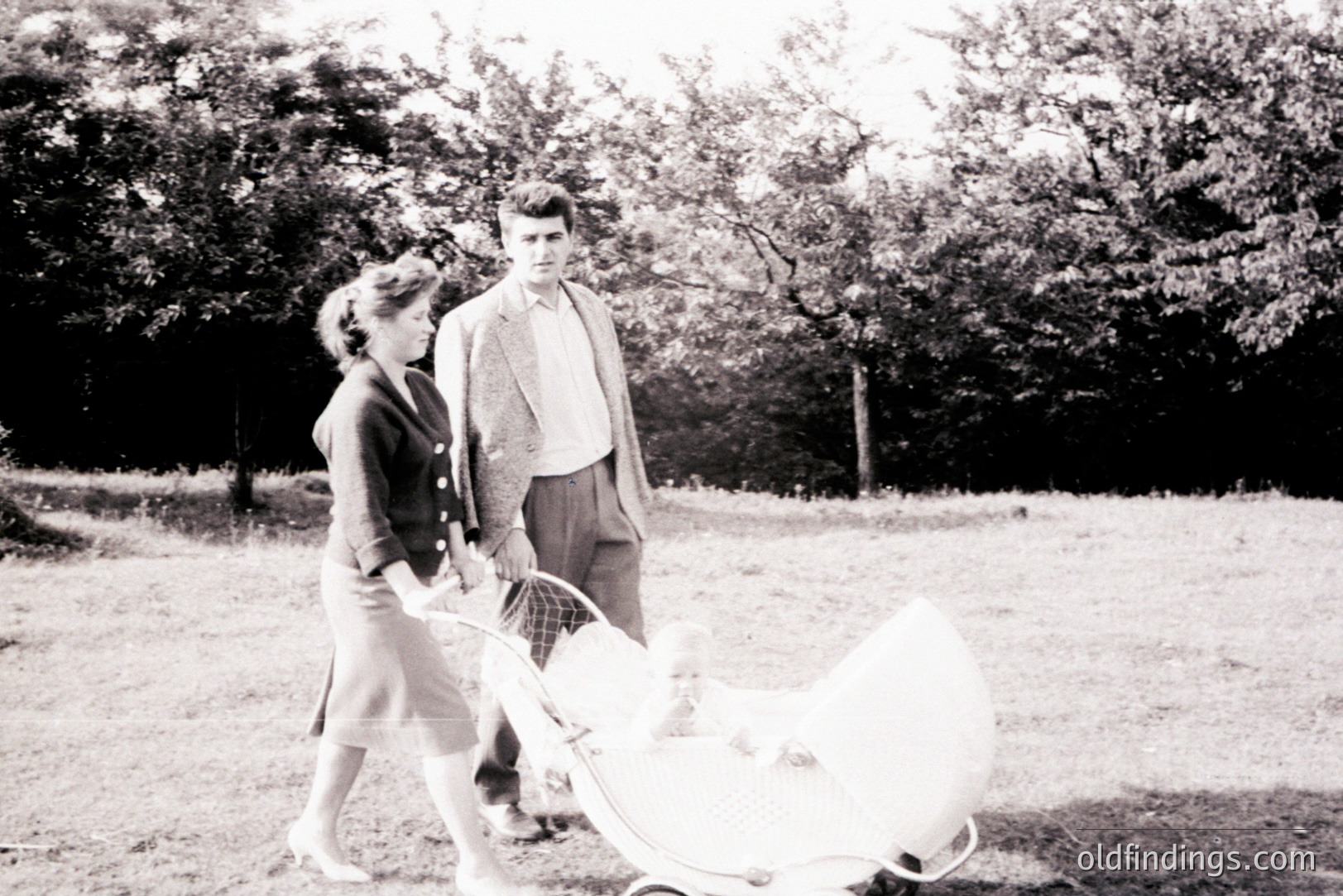 A young couple strolls through a grassy area, pushing a vintage wicker baby carriage. The woman wears a tailored suit, and the man a tweed jacket; attire typical of the 1950s. Soft lighting & simple composition suggest a candid family moment.