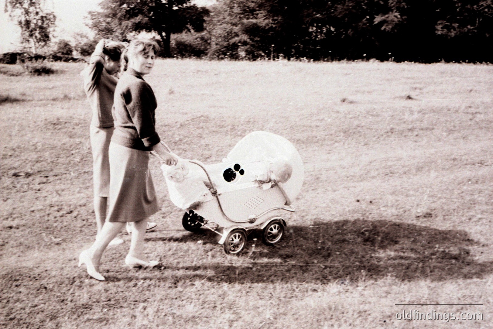 A woman in a 1960s-style dress and heels pushes a vintage, ornate baby carriage across a grassy field. Another figure is partially visible, walking alongside. Simple, natural background. Evokes domesticity & mid-century design. Potential for stock imagery, design references.