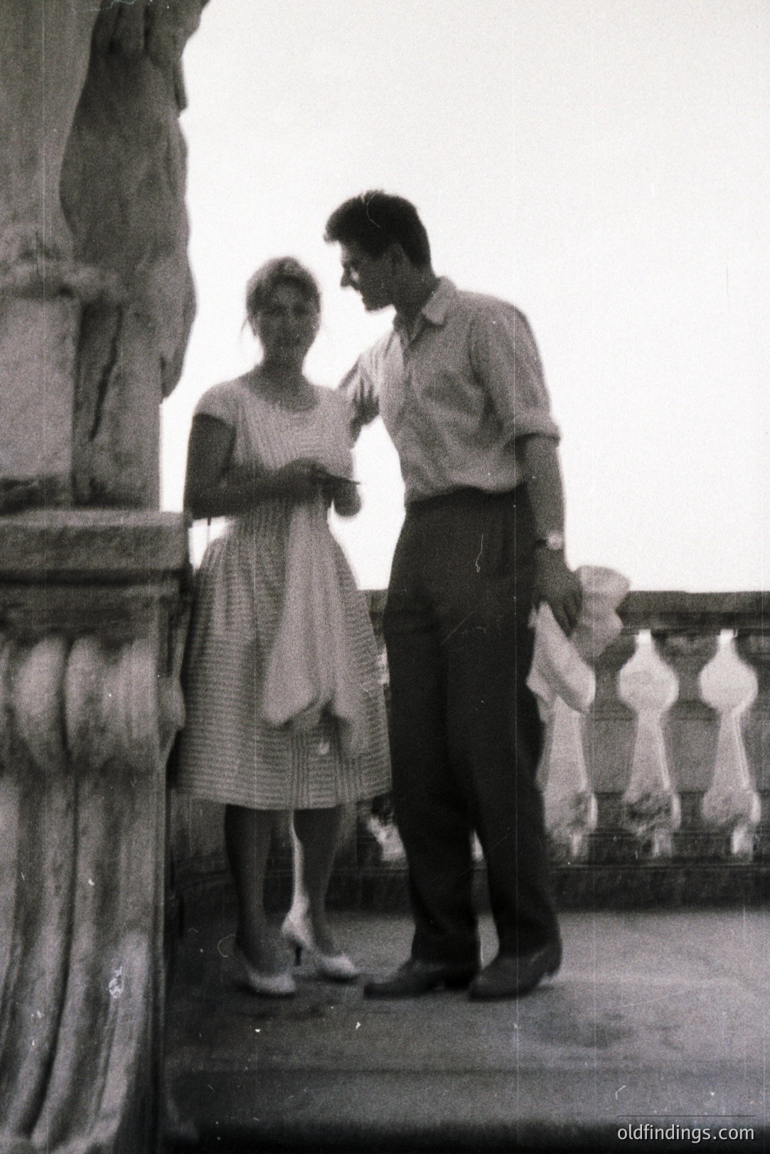 A stylish couple poses near an ornate stone balustrade. The woman wears a patterned dress & heels; the man, a short-sleeved shirt & trousers. Likely a travel photo from the 1960s, depicting a leisurely moment. Architectural detail suggests a European location. Strong potential for retro design and historical research use.