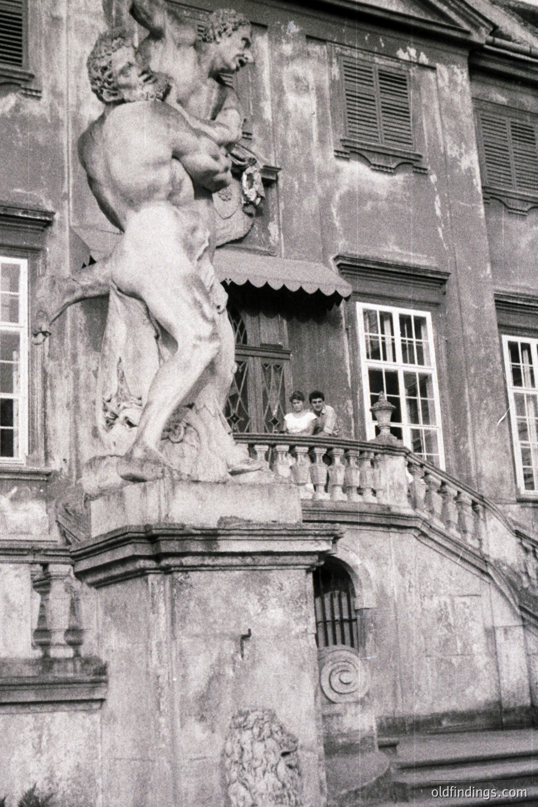 Striking monochrome image showcases a weathered stone sculpture of a muscular man, possibly a mythological figure, atop a monumental stone facade. Two figures are visible on a balcony above. Likely late 19th or early 20th century. Captures architectural detail and subtle human presence.