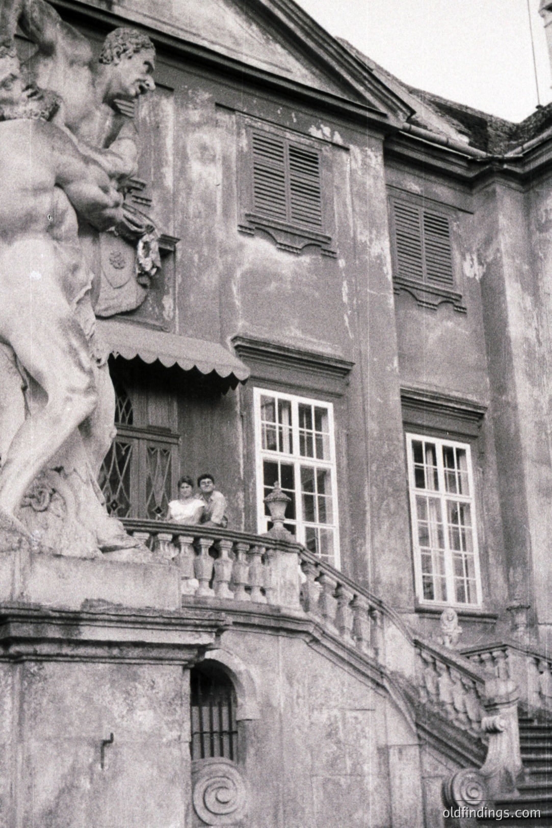 Elegant, aging estate facade with a sculpted figure overlooking tiered steps and a balcony where two figures stand. Building features shuttered windows and a classically-inspired design. Likely a formal garden or villa setting, possibly European. Appears to be a vintage print, possibly 1930s-1960s.