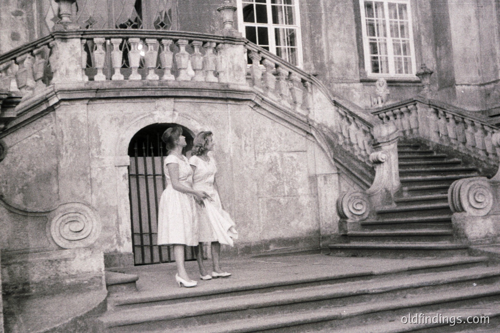 Two young women in matching knee-length dresses and heels pose on a grand stone staircase. The ornate architecture features a balustrade, decorative arches, and a large, imposing gate. Likely a formal garden or estate, possibly European. Estimated timeframe: 1960s. Suitable for design and historical reference.