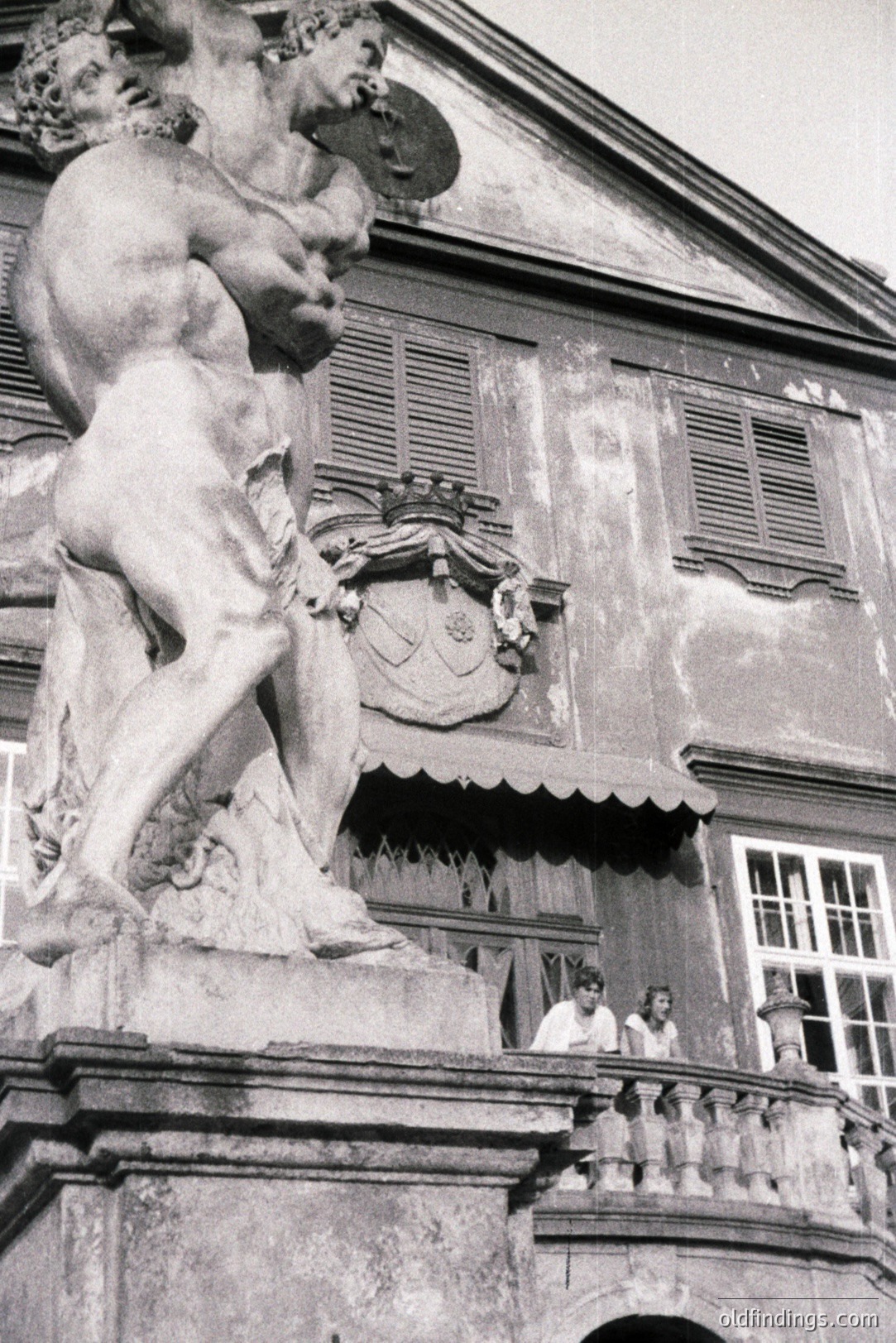 Monumental marble statue of a muscular figure, likely a mythological subject, stands prominently before a weathered, shuttered facade. Two women observe from a balcony above, hinting at a stately estate setting. Appears to be a formal garden or courtyard scene.