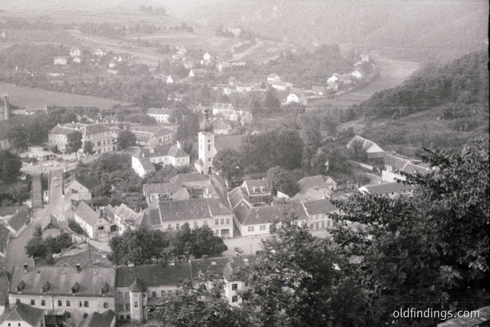High-angle view of a European town, likely Central or Eastern Europe, featuring tightly-packed buildings with gabled roofs clustered around a central church spire. The landscape shows rolling hills and forested slopes. Appears to be a late 19th or early 20th-century photograph.