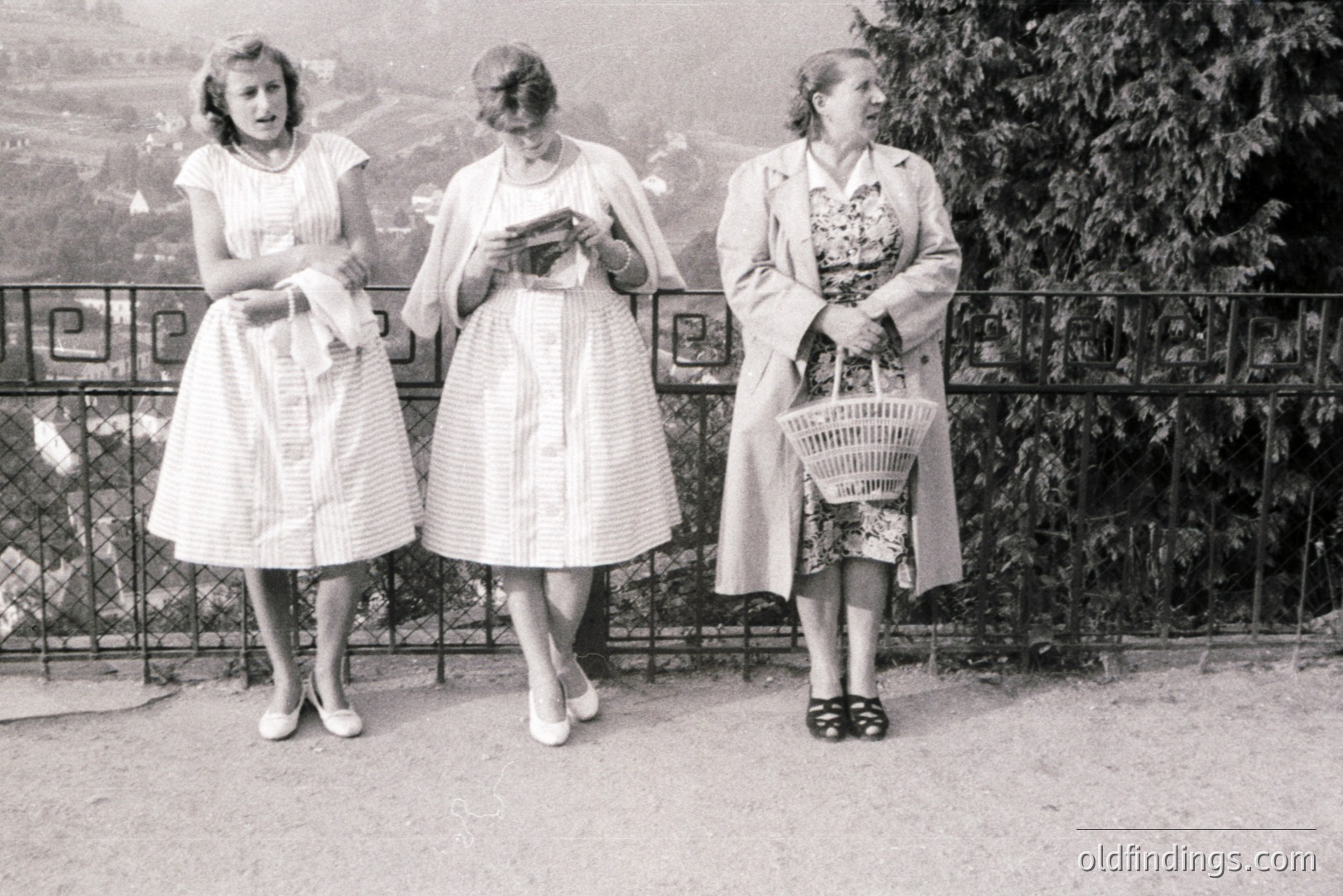 Three women stand by a wrought-iron railing overlooking a landscape. All wear patterned summer dresses and carry accessories: one holds gloves, one a book, and the third a woven basket. Likely 1950s, possibly European, showcasing mid-century fashion and leisure. The image has potential for design or historical research.