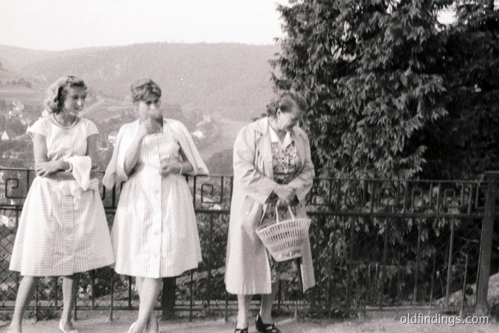 Three women in 1950s dresses and outerwear observe a scenic view from a garden terrace. Wrought-iron railings and lush greenery frame the background. One woman carries a woven basket. Likely a candid snapshot documenting leisure time.