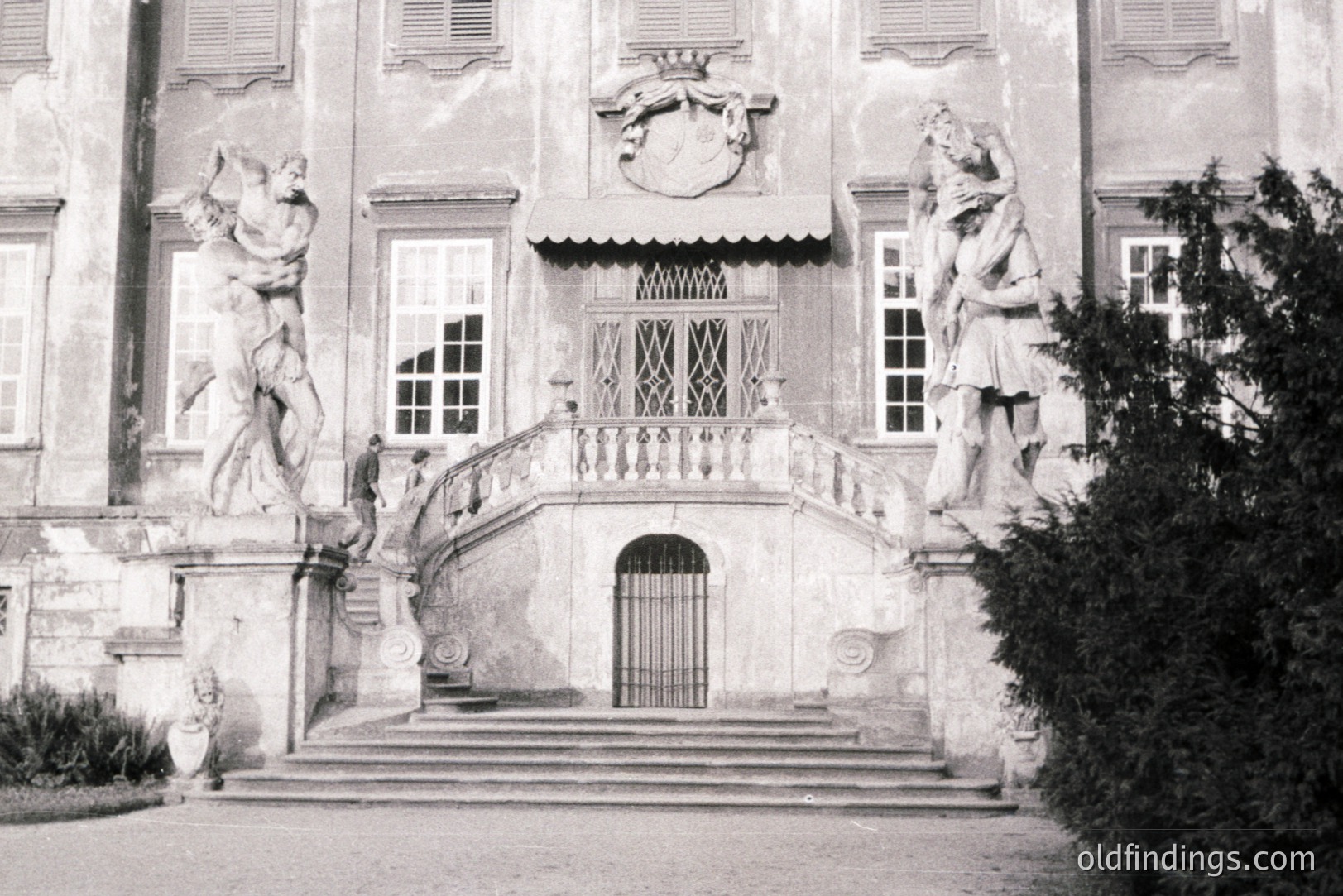 Formal, symmetrical architecture features a central entrance flanked by dramatic allegorical statues. Stone steps lead to an ornate doorway, showcasing classical design elements & a decorative crest above. Likely an estate or palace.