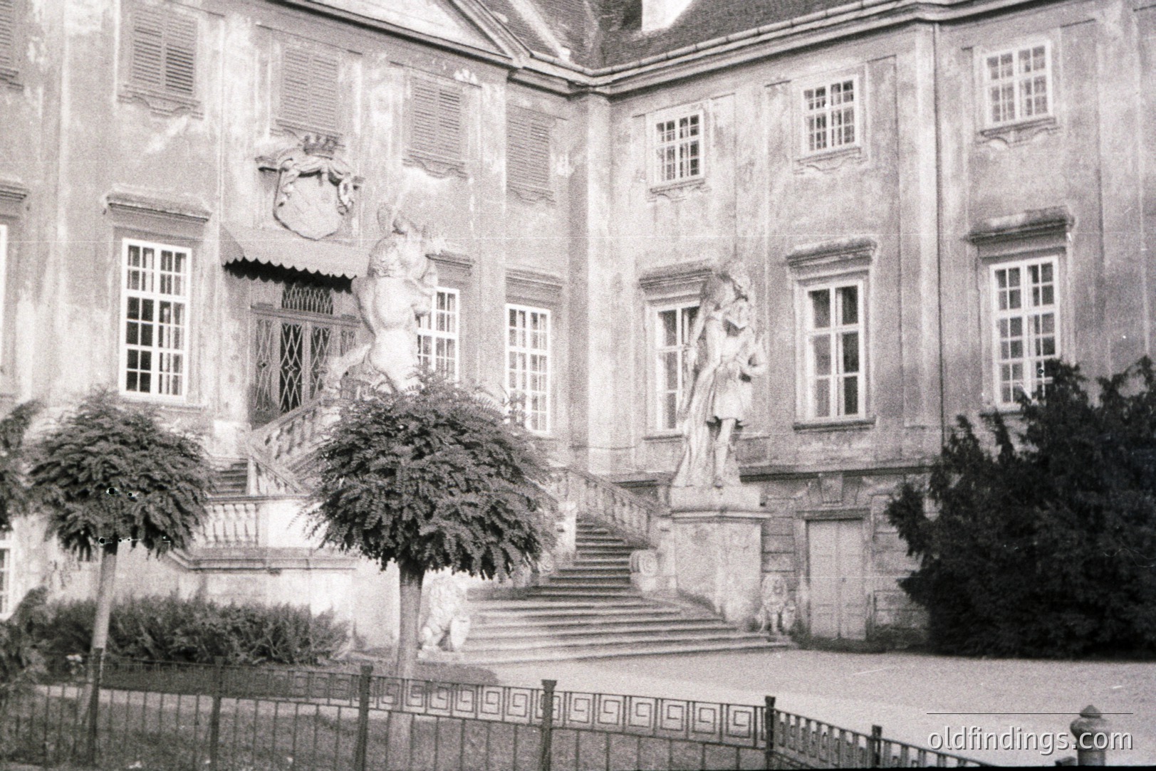 Ornate stone staircase leads to a monumental building façade featuring symmetrical windows and a heraldic crest. Formal topiary gardens frame the entrance. Likely an aristocratic estate, possibly 18th-century European design. Could be useful for architectural reference or historical context.