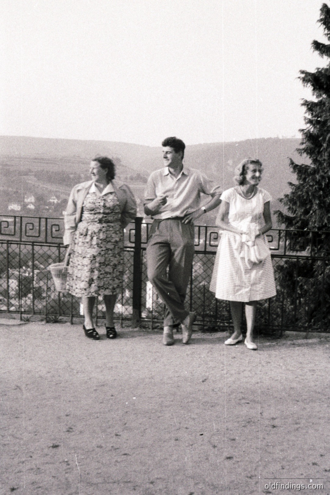 Three figures pose outdoors along a railing with a hillside view. The woman on the left carries a wicker basket. The man wears a short-sleeved shirt, and the woman on the right has her arm raised. Likely 1950s or 60s attire and composition. Scenic landscape.