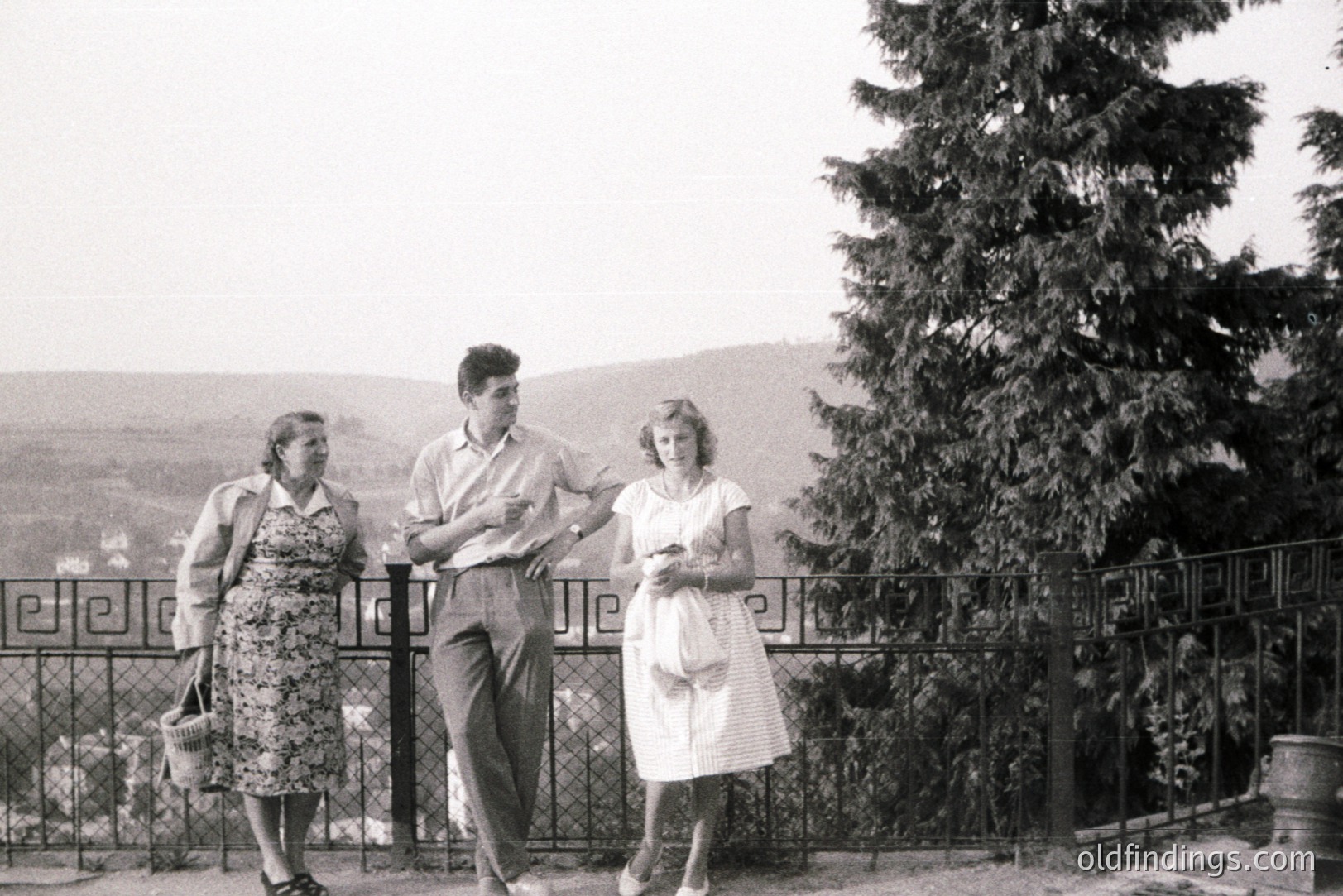 A relaxed group portrait: A woman in a dress and jacket, a young man in slacks & short-sleeved shirt, and a woman in a floral dress gesture towards a hillside view. Likely 1960s fashion. Possible seaside resort setting with wrought iron fence. Appealing for design/stock usage.