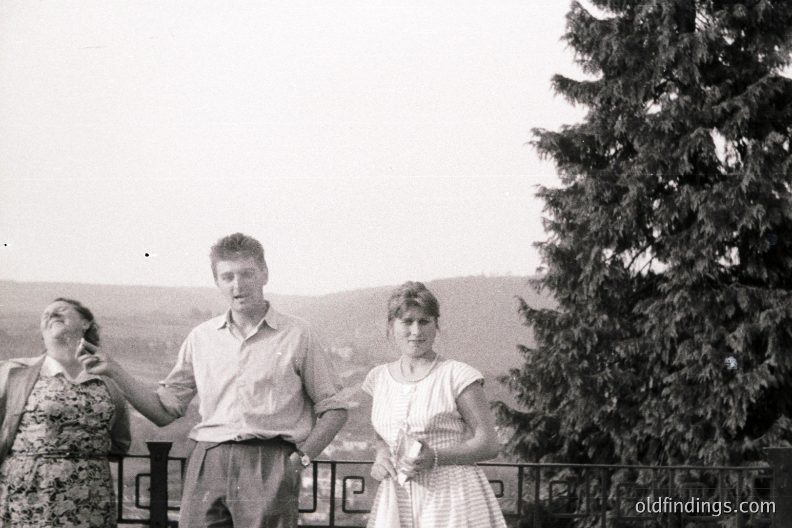Three young adults stand on a balcony overlooking a landscape with a large pine tree dominating the right side. The man is centered, flanked by a woman on the left and another on the right. Clothing suggests the 1970s - 1980s. Likely a vacation snapshot.