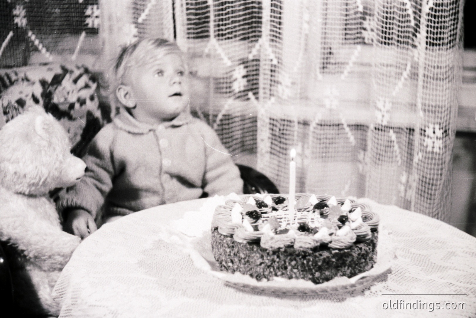 A young child, seated in a chair, gazes upward at a birthday cake adorned with berries and a single lit candle. A teddy bear rests beside the child. The scene, likely a domestic setting, is framed by a lace-curtained window. Appears to be a candid moment, likely 1960s or 70s.