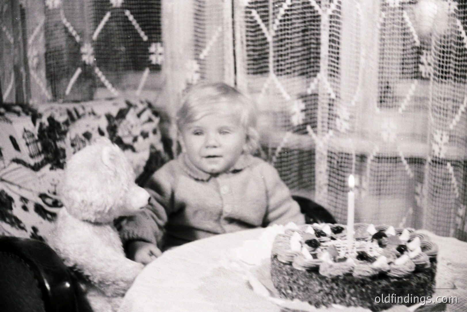 A young child sits next to a large teddy bear and a birthday cake with lit candles. The scene is framed by patterned curtains and a floral armchair. Likely a family snapshot from the 1960s or 70s, capturing a moment of childhood celebration. Nostalgic, domestic imagery.