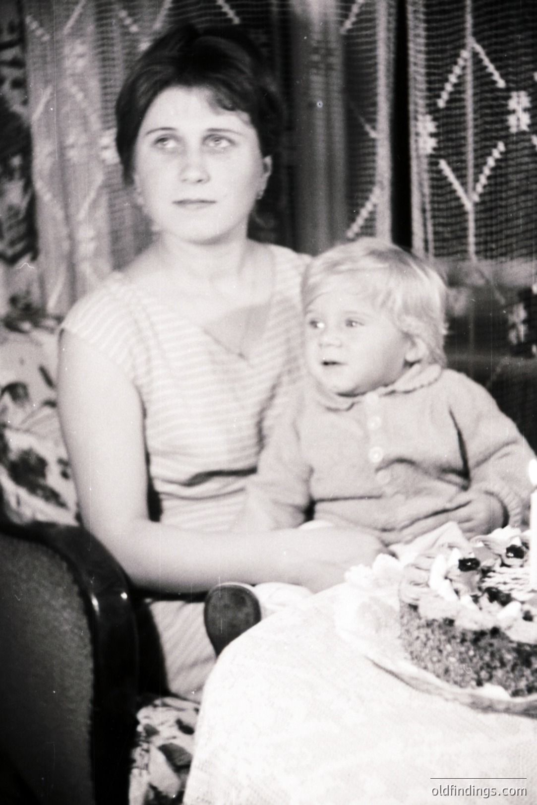 A seated woman holds a young child, both appearing to gaze directly at the camera. A decorated cake with lit candles sits on a table to the right. The scene suggests a birthday celebration, likely within a domestic interior. Style and clothing indicate a mid-1960s timeframe. Photo’s texture and lighting are hallmarks of vintage film photography.