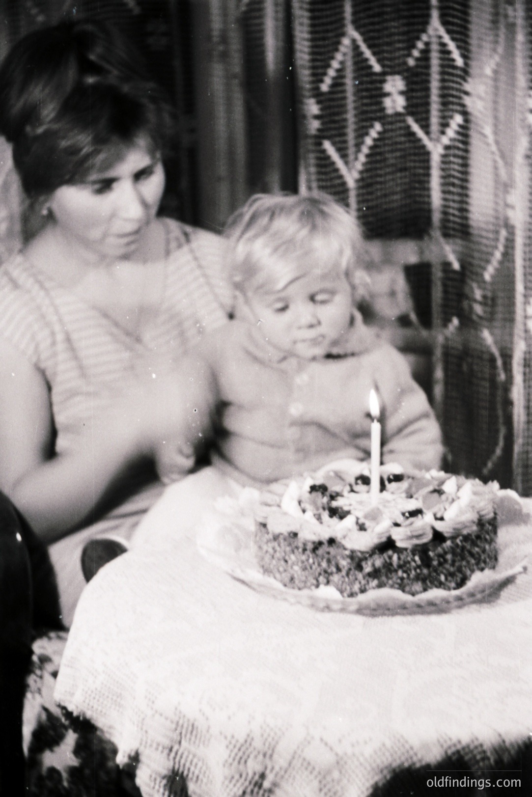 A candid moment captures a young child seated before a cake adorned with cherries and a single candle. The child appears contemplative, possibly anticipating a wish. A woman's hand gestures nearby, likely assisting the child. Likely a family gathering, possibly a birthday celebration. Appears to be a 1960s or 70s snapshot.