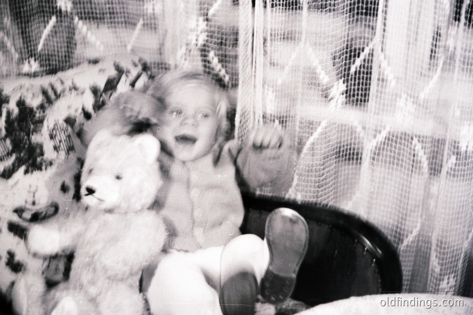 Young child seated in a high chair, clutching a fluffy teddy bear. Likely a family snapshot, showcasing a moment of childhood play. The background features decorative lace, suggesting an interior setting. Appears to be a candid, informal portrait. 1960s-1970s timeframe.