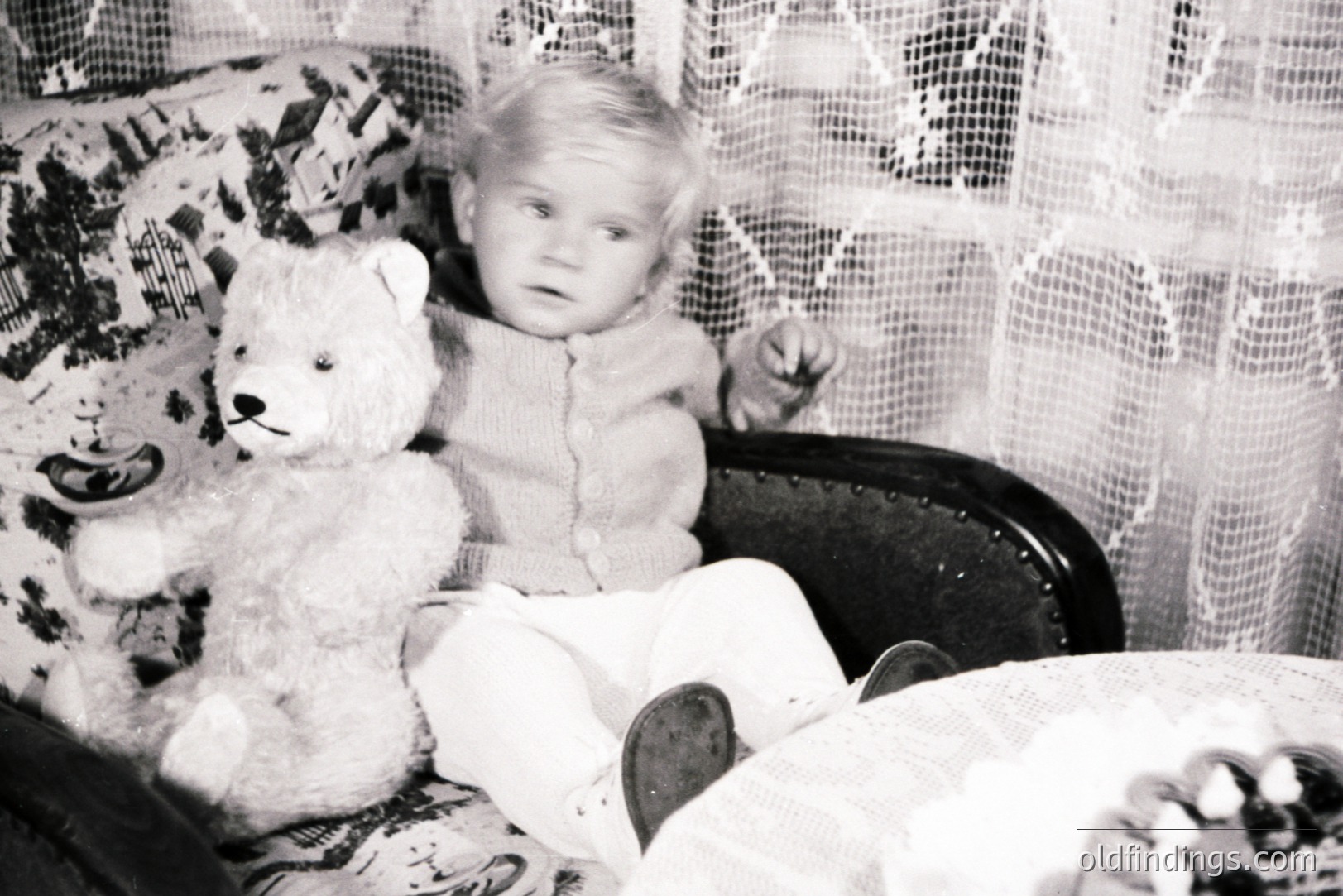 Young child seated in an ornate chair, clutching a worn teddy bear. Dressed in a knitted sweater and trousers, posed formally. Detailed lace backdrop suggests a domestic interior. Likely a family portrait from the 1940s-1960s. Potential for sentimental value, vintage design reference.