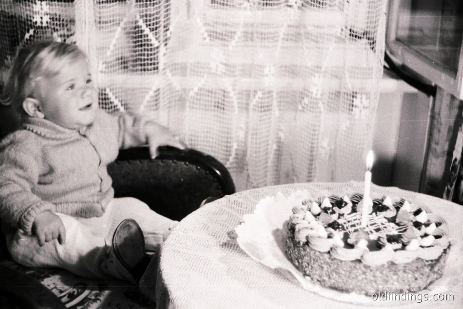 A young child sits in a dark wicker chair, gazing at a layered cake adorned with cream frosting and a single lit candle. The scene, framed by lace curtains, evokes a domestic, celebratory moment. Likely a family snapshot from the 1960s or 1970s. Appears in vintage, black and white.
