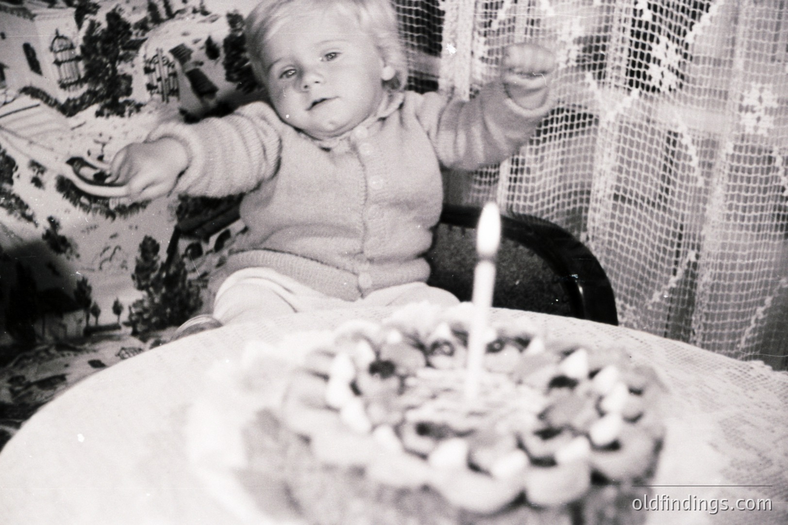 A young child sits in a highchair, reaching for a frosted cake with a single lit candle. The setting appears to be a domestic interior with patterned wallpaper and lace curtains. Likely 1960s-70s snapshot capturing a childhood milestone. Commercial value as vintage family life reference.