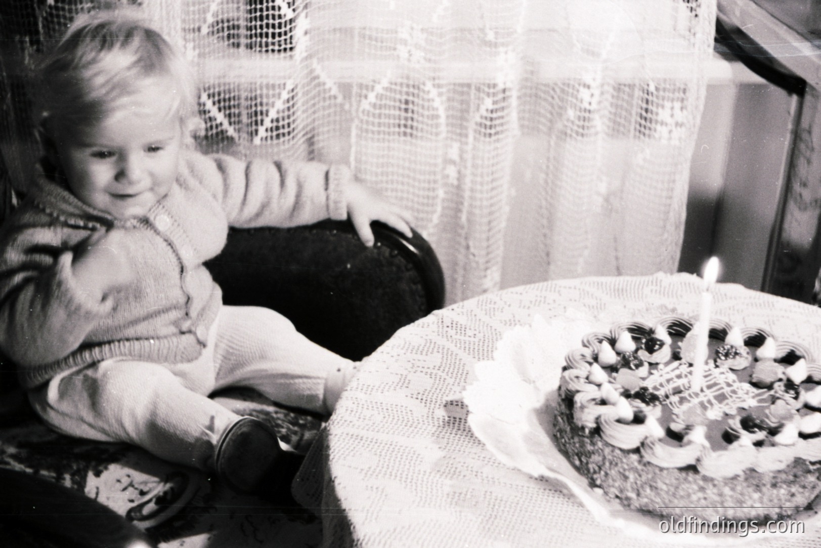 A young child, likely a toddler, sits in a cushioned chair, observing a decorated cake with a single lit candle. The scene suggests a birthday celebration, captured in a classic black and white photograph. Likely 1950s-1960s, domestic interior with lace curtains. Image has archival value, potential for nostalgia-themed design.