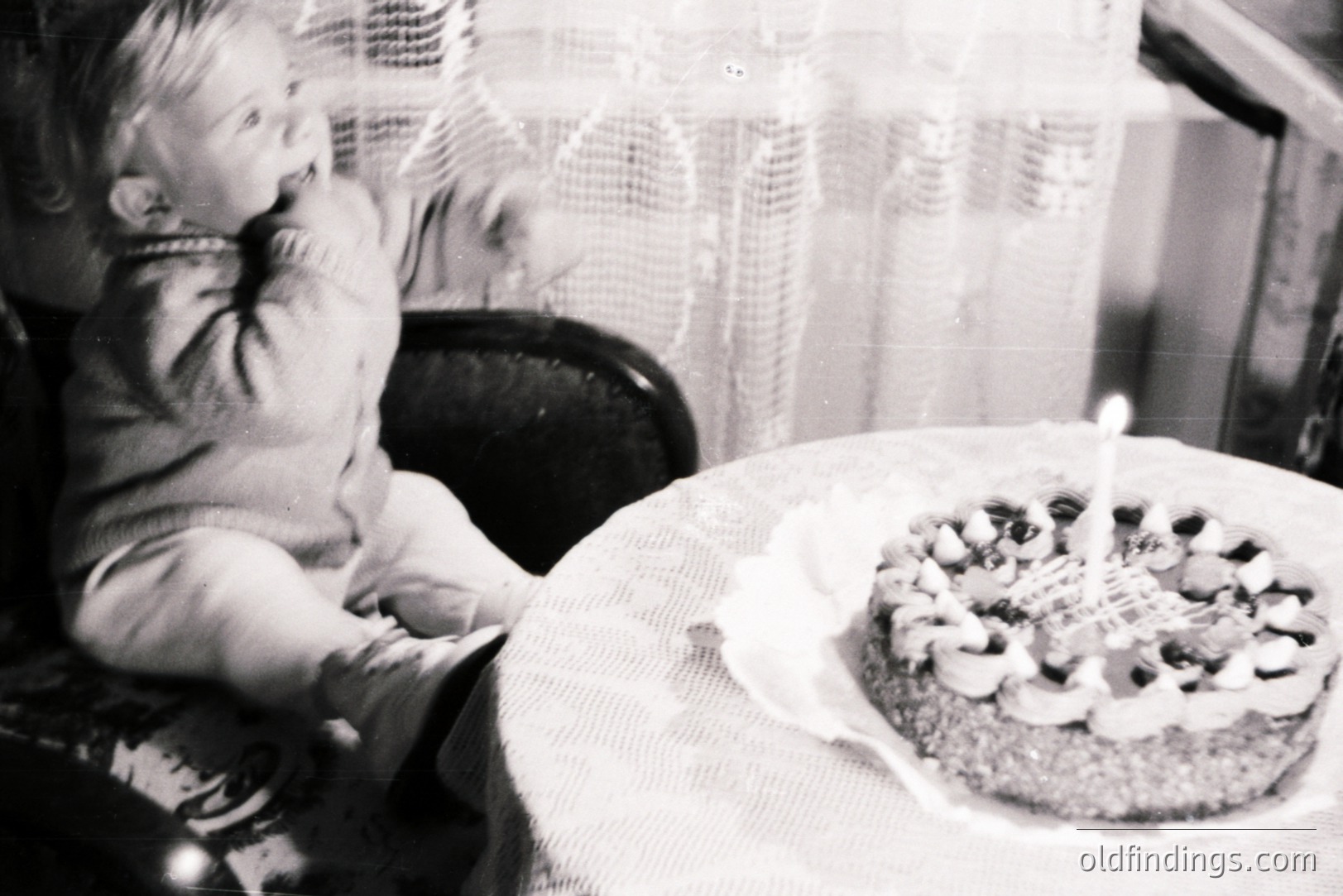 A young child, likely a toddler, sits in a high chair, reaching towards a decorated cake with a single lit candle. The cake sits on a lace-covered table. The image suggests a celebratory, intimate moment, likely a birthday. Appears to be from the 1950s or 60s.