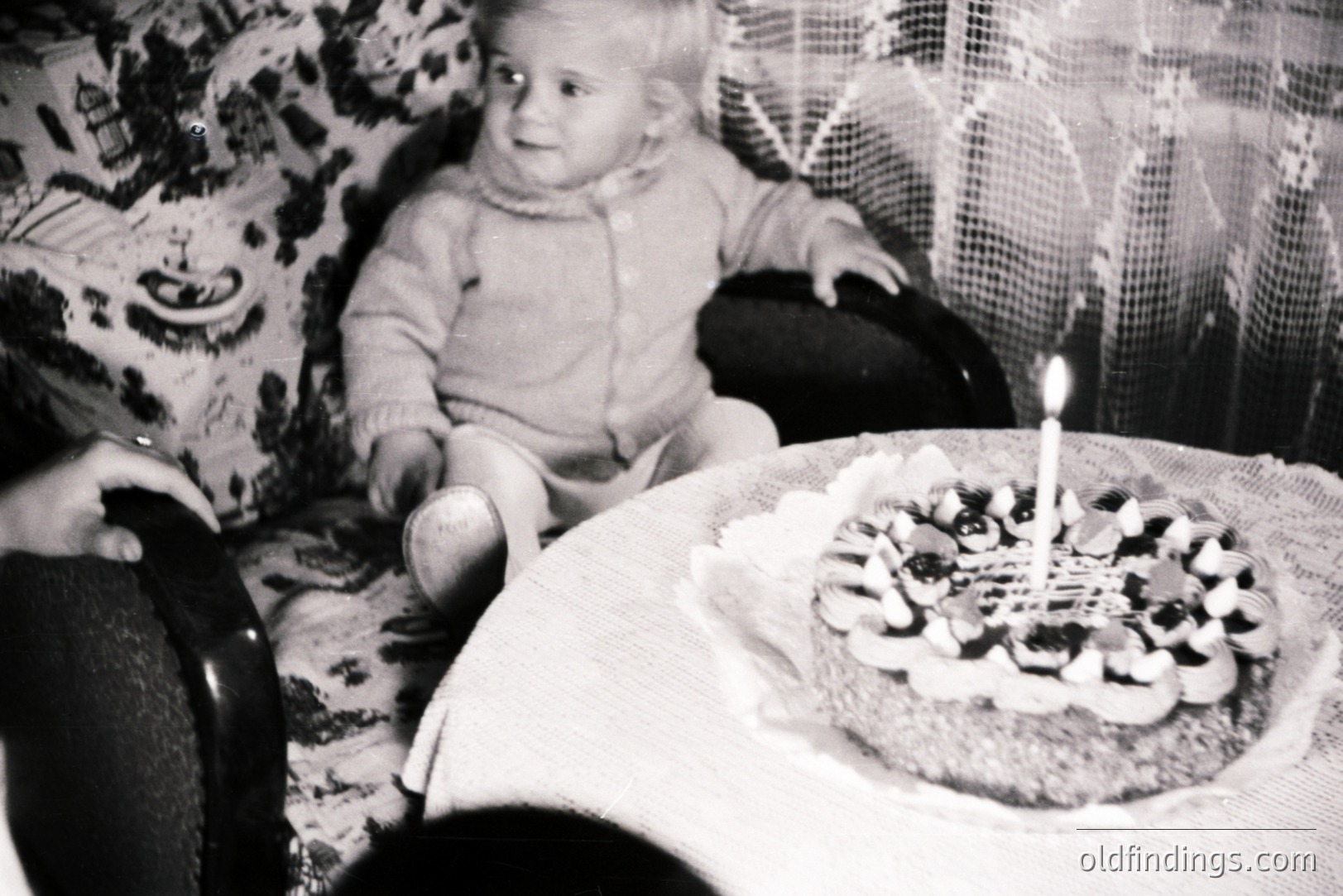 A young child, possibly a toddler, sits in an ornate armchair, appearing to observe a cake decorated with what appear to be sugared almonds. The scene suggests a celebration, likely a birthday. A lace-covered table holds the cake. Appears to be a family snapshot, circa 1960s.