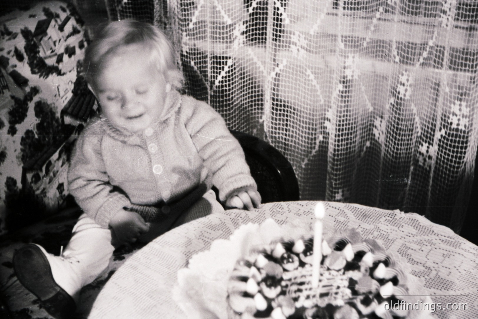 A young child, seated at a table covered with a lace tablecloth, gazes towards a small cake adorned with a single lit candle. The scene, captured in monochrome, evokes a nostalgic, intimate atmosphere. The child wears a textured knit sweater. Likely a family snapshot, circa 1960s-1970s.