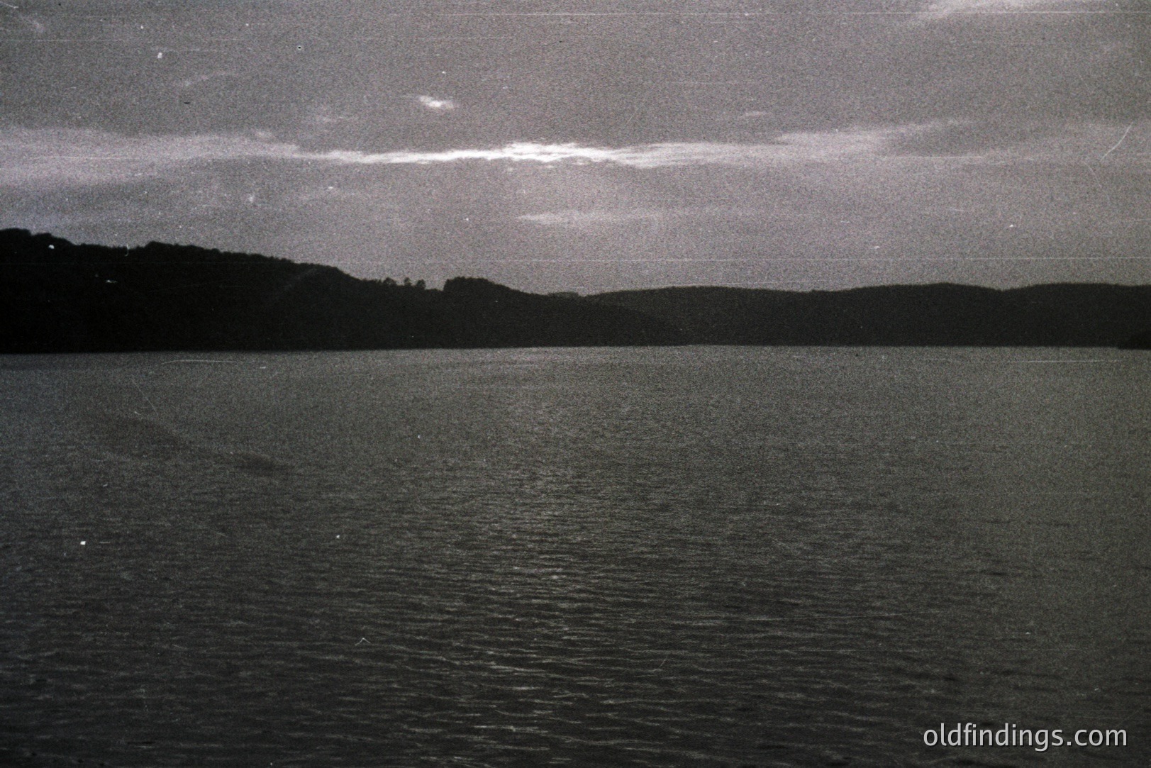 Moody black and white landscape with a still body of water reflecting an overcast sky. A dark shoreline and dense trees define the horizon line. Visible grain suggests film photography. Possibly a coastal scene. Atmospheric and evocative.