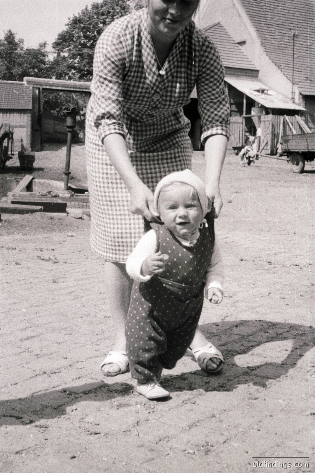 A young child in a polka-dotted overalls and bonnet takes tentative steps while being supported by an adult's hand. Farmhouse exterior visible, with rustic tools and a weathered wheelbarrow in the background. Likely mid-20th century, possibly rural Europe. A charming domestic scene depicting early childhood.