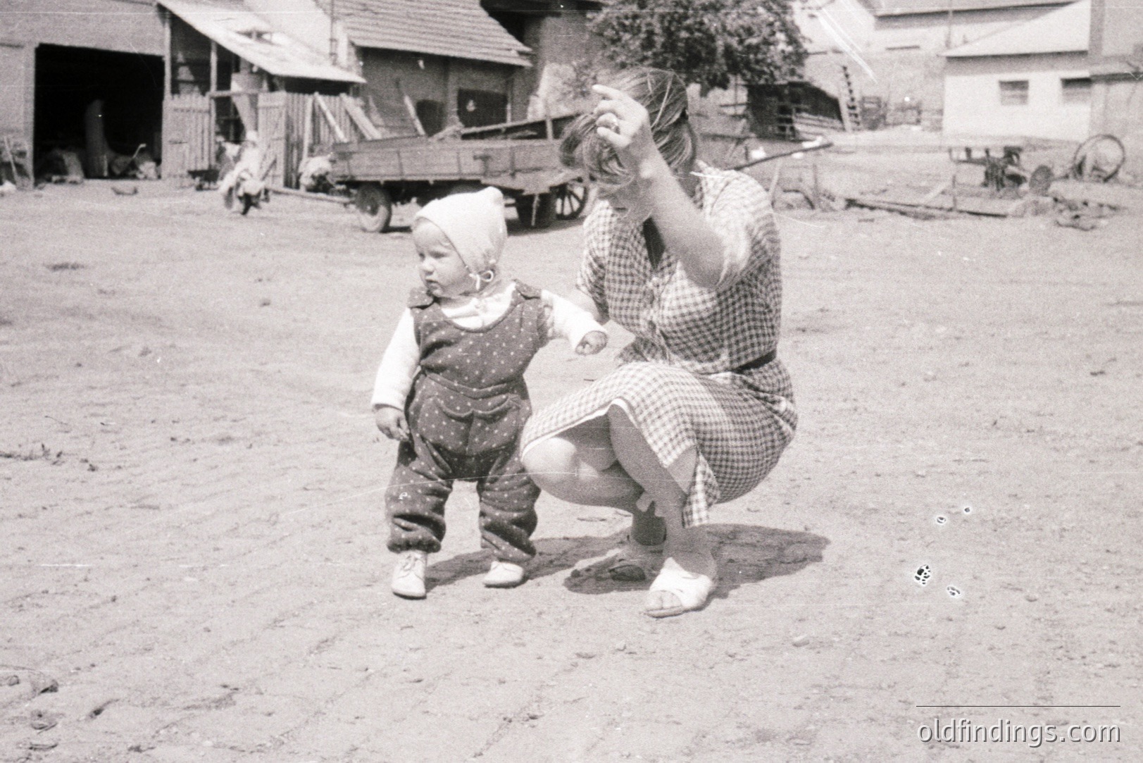 A young child in overalls and cap takes tentative steps outdoors. A woman in a checked dress squats, blowing bubbles. Rural setting features a farmhouse, wooden cart, and outdoor workspace. Likely a family snapshot, c. 1950s-1960s. Documents daily life and family dynamics.