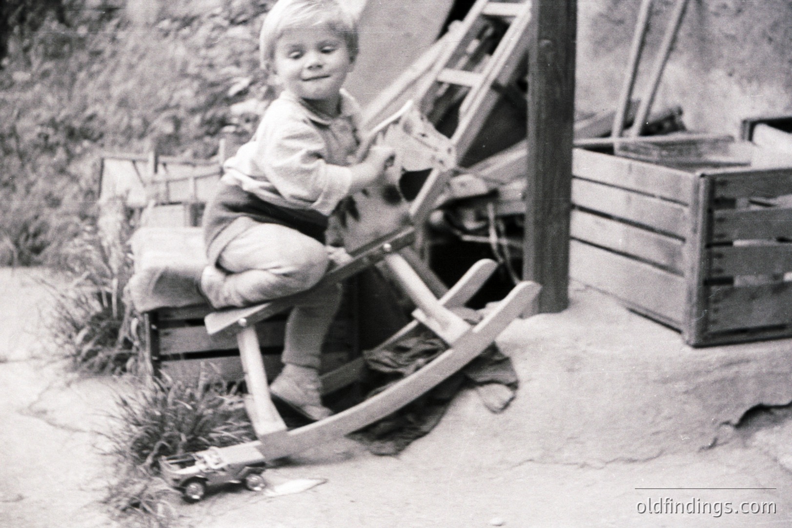 A young boy sits on a wooden rocking horse, smiling. He wears overalls and a collared shirt, suggesting a mid-20th century, likely 1950s setting. A toy truck sits nearby on a sandy ground. Rustic wooden boxes and a ladder add to the scene. Simple, nostalgic childhood moment.