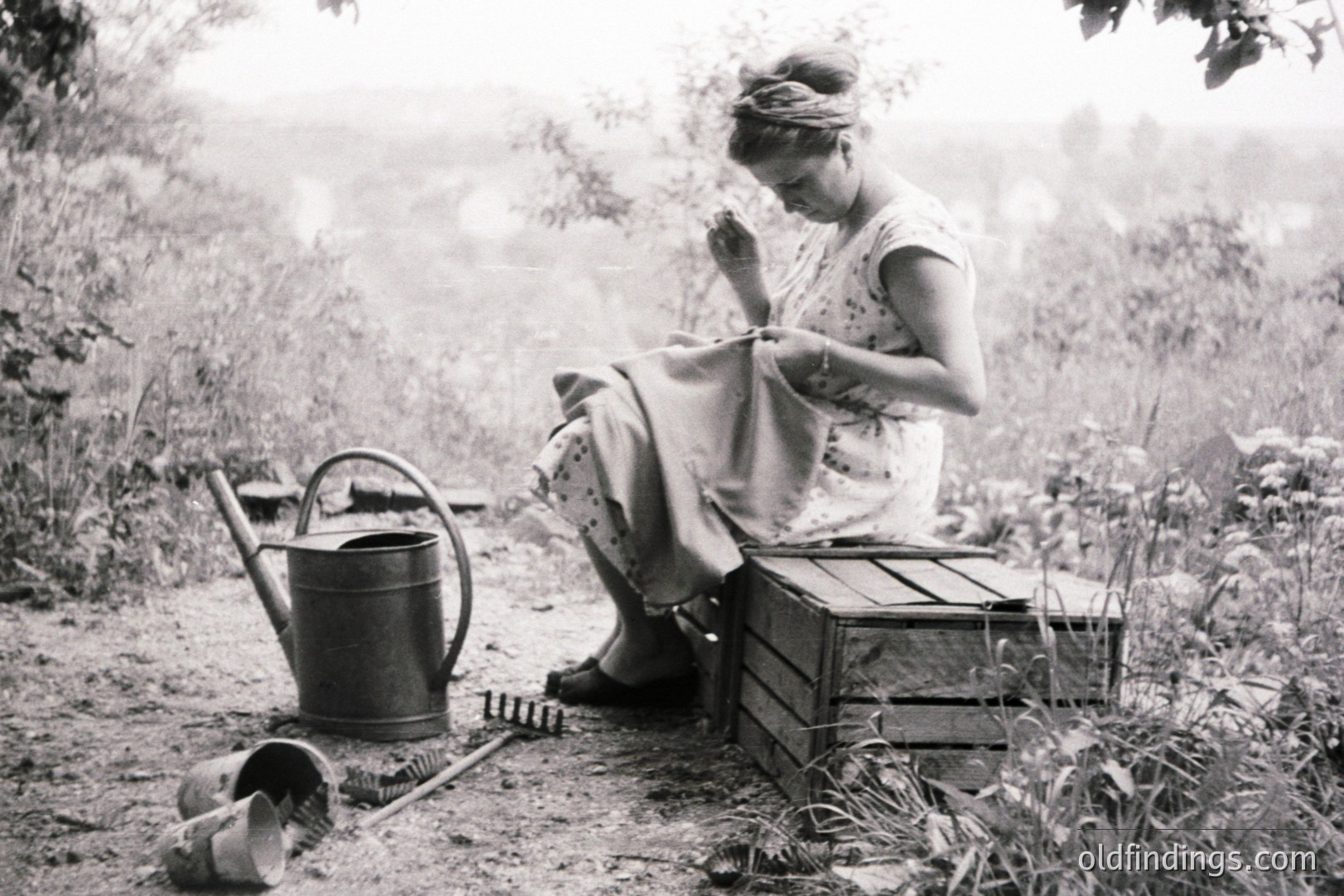 A woman seated on a wooden crate, focused on mending fabric with a needle. Gardening tools, including a watering can and small trowels, suggest a domestic or rural setting. Likely a mid-20th century scene, capturing a moment of quiet industry.