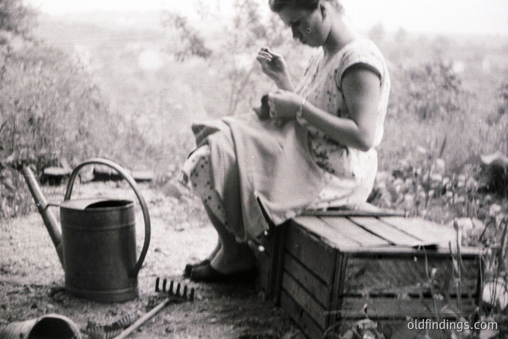 A woman sits outdoors, engaged in needlework on a wooden crate. A watering can and gardening tools rest nearby, suggesting a rural setting. Likely mid-20th century, the image holds nostalgic charm & potential for design use. The photograph's textures evoke a simple, domestic life.
