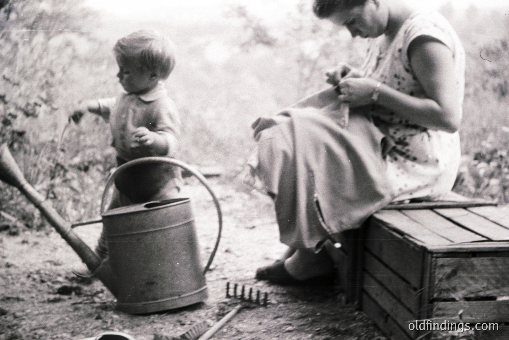 A young boy stands near a watering can, while an older woman sits on a wooden crate, seemingly engaged in needlework. Rustic garden tools rest nearby, suggesting outdoor labor. Likely a candid family moment from the mid-20th century, evoking a sense of domesticity and rural life.
