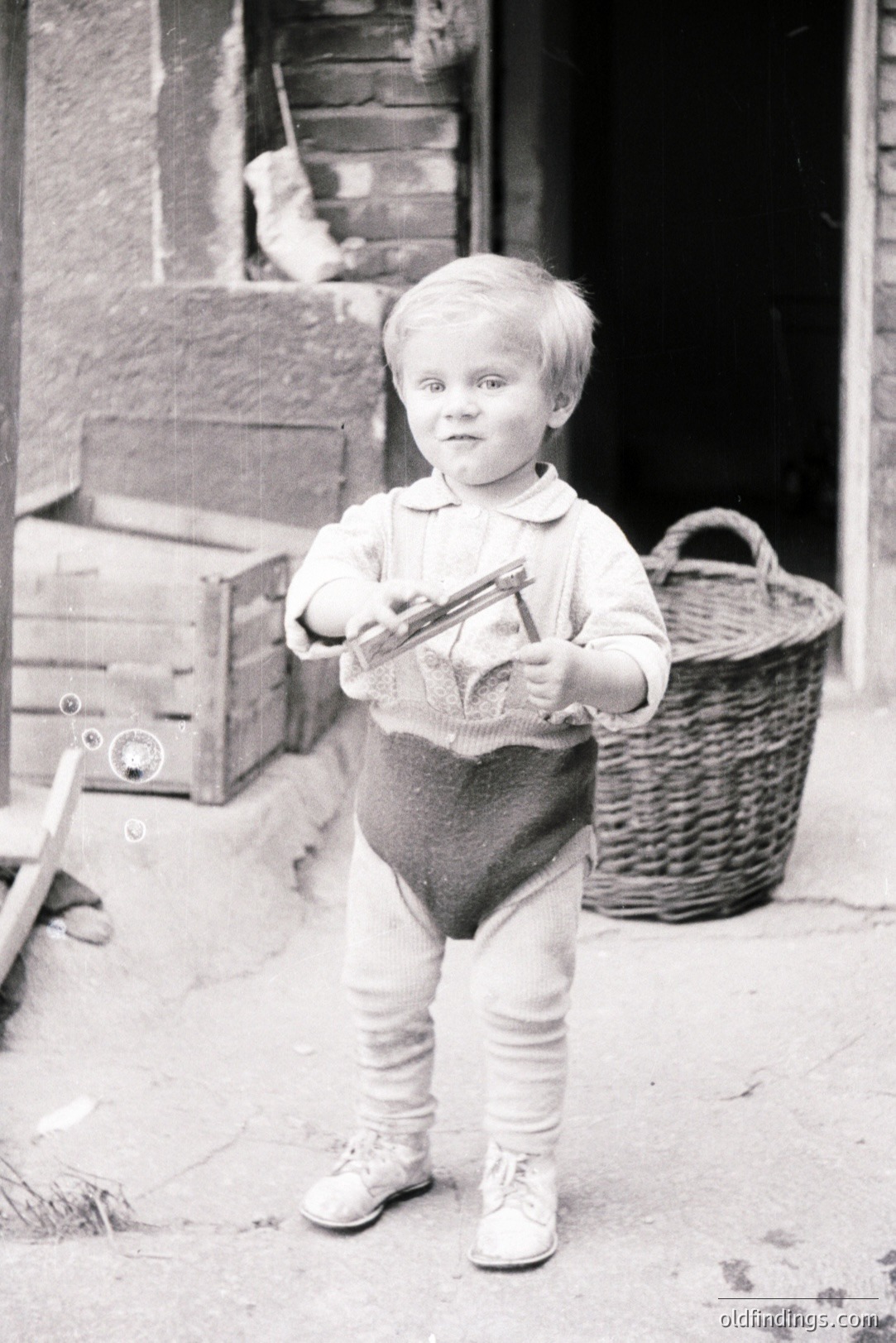 A young boy, standing outdoors, holds a small, antique-style camera. He's dressed in a collared shirt, bloomers, and shoes. A woven basket and stacked crates are visible behind. Likely early-mid 20th century, this photo evokes childhood and nostalgia. A small detail - bubbles in the foreground.