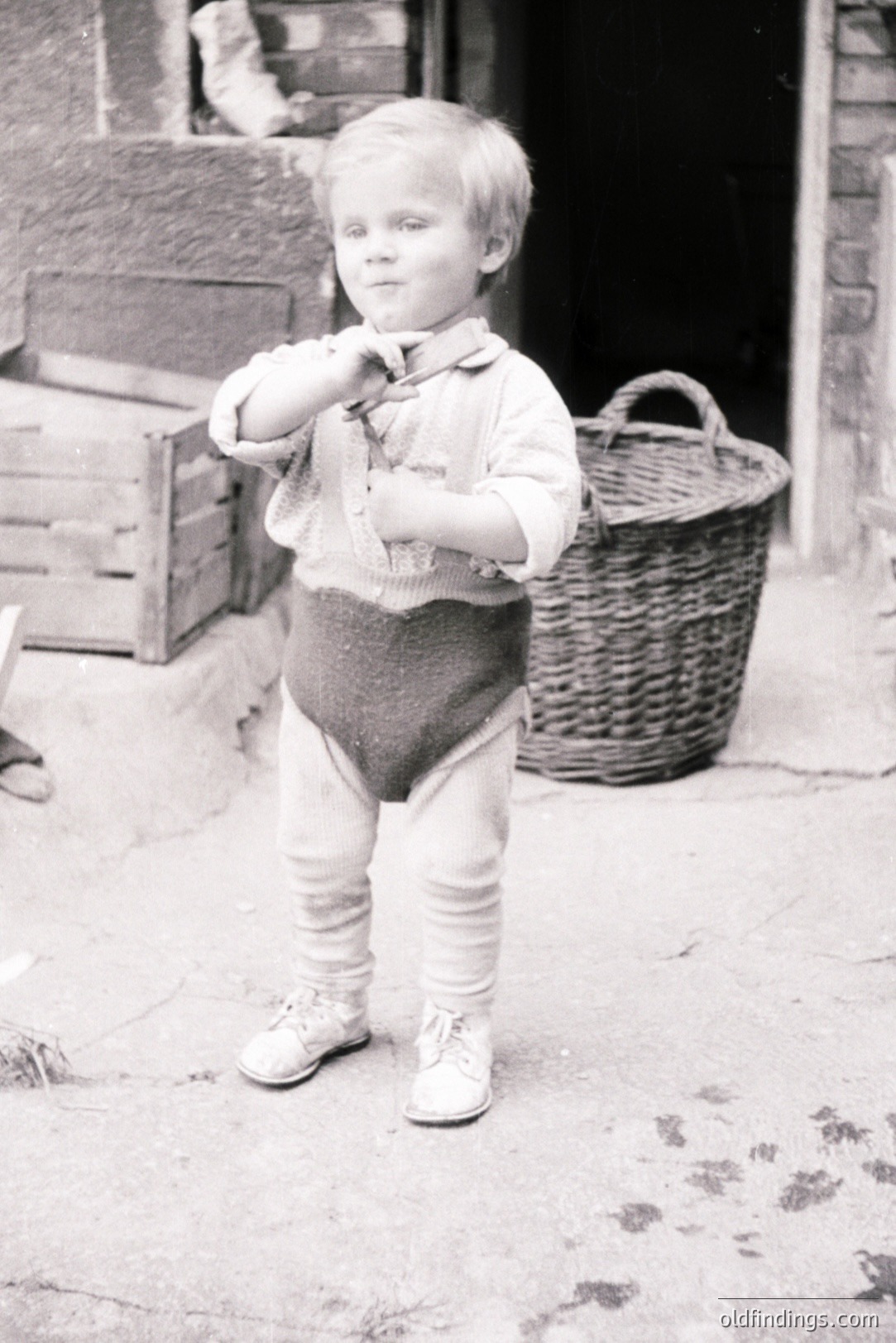 A young boy stands outside a building, wearing knit stockings, shorts, and a collared shirt. He poses with a playful expression, slightly puckering his lips. A wicker basket and stacked crates are visible nearby. Likely a candid portrait, c. 1930s-1950s.