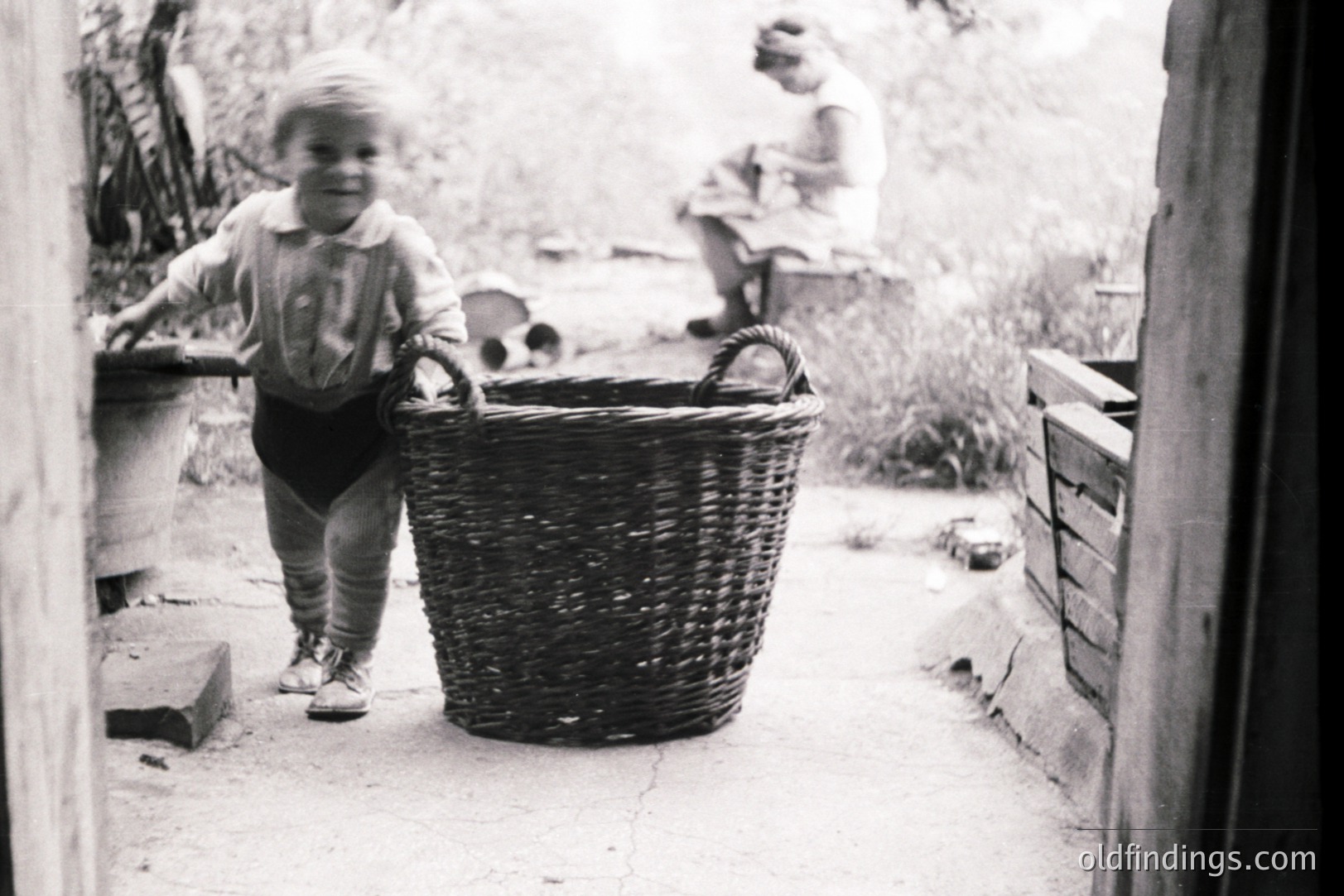 A young child in short overalls stands beside a large, woven basket, seemingly offering it. A woman, wearing a light-colored dress and head covering, is seated nearby on a rough-hewn stool. The scene appears to be a rural yard setting, framed by a doorway. Likely 1930s-1950s. Photographic style is characteristic of documentary or family snapshots.