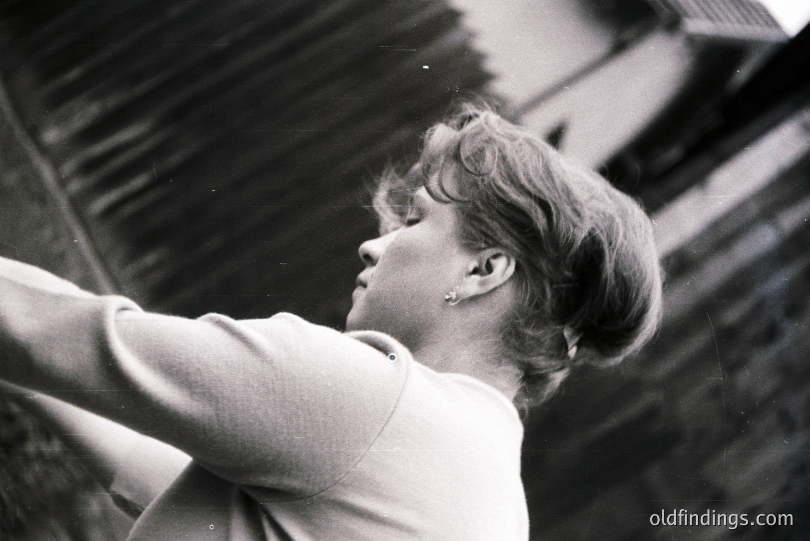 A young woman with short, styled hair is captured in profile, arms raised, against a weathered wooden building facade. The image shows subtle grain and wear characteristic of older film stock. Likely a candid portrait, possibly mid-1960s or 70s. A simple earring is visible.