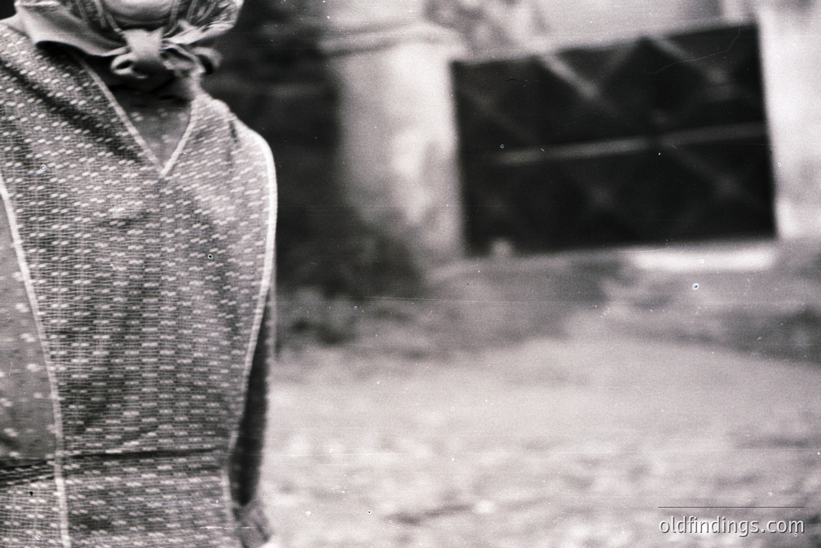 A detail view of a person wearing a patterned jacket and headscarf. The subject stands facing away from the camera, partially obscured by foliage, before a blurred, rustic stone building. Likely a candid snapshot, 1960s or 70s aesthetic. Evokes vintage fashion and documentary style.