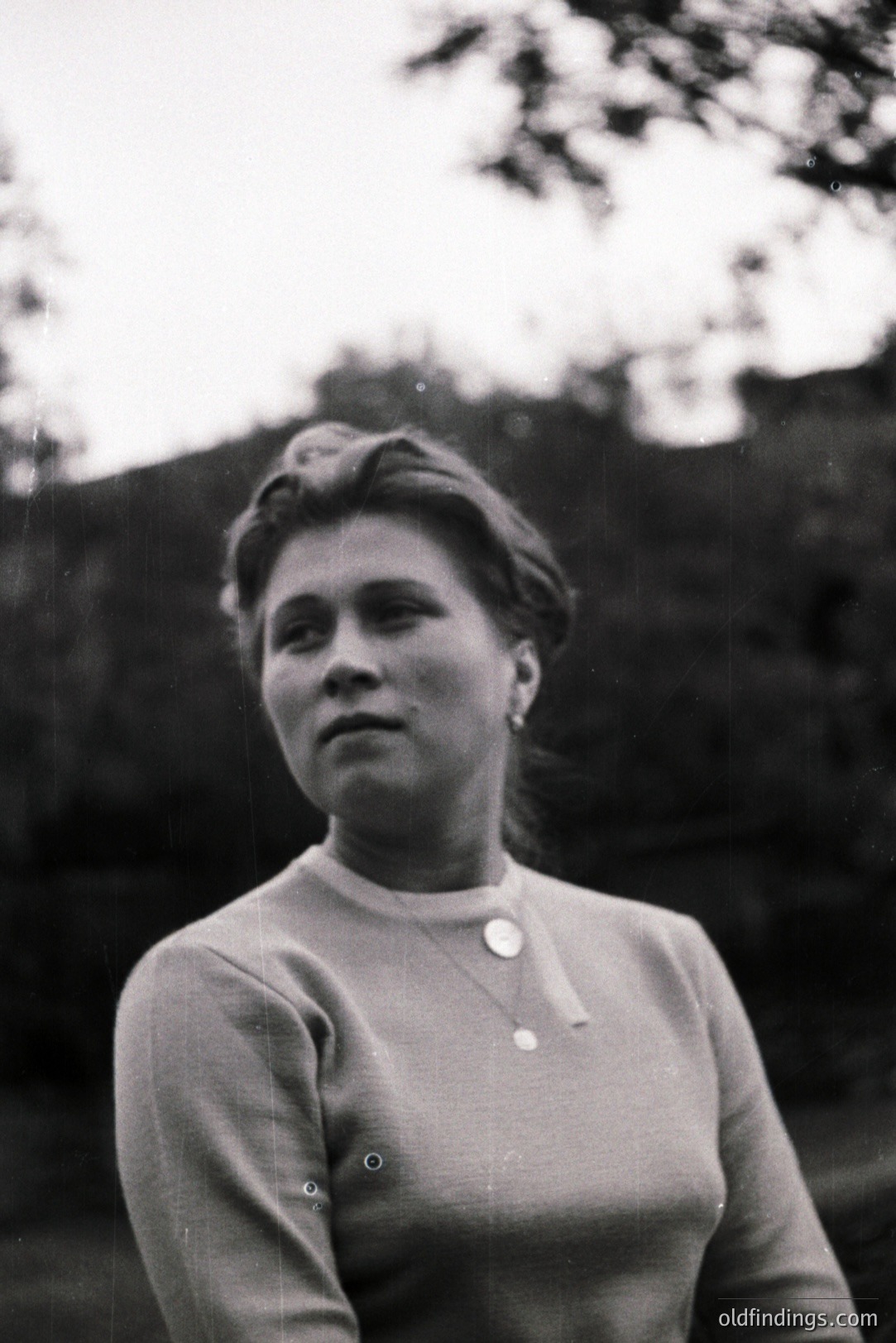 A portrait of a woman in a high-necked sweater, likely 1960s fashion, with a simple pendant necklace. Soft lighting and shallow depth of field create a focus on her face, framed by foliage. A casual, posed outdoor setting. Appears suitable for vintage design or family history research.