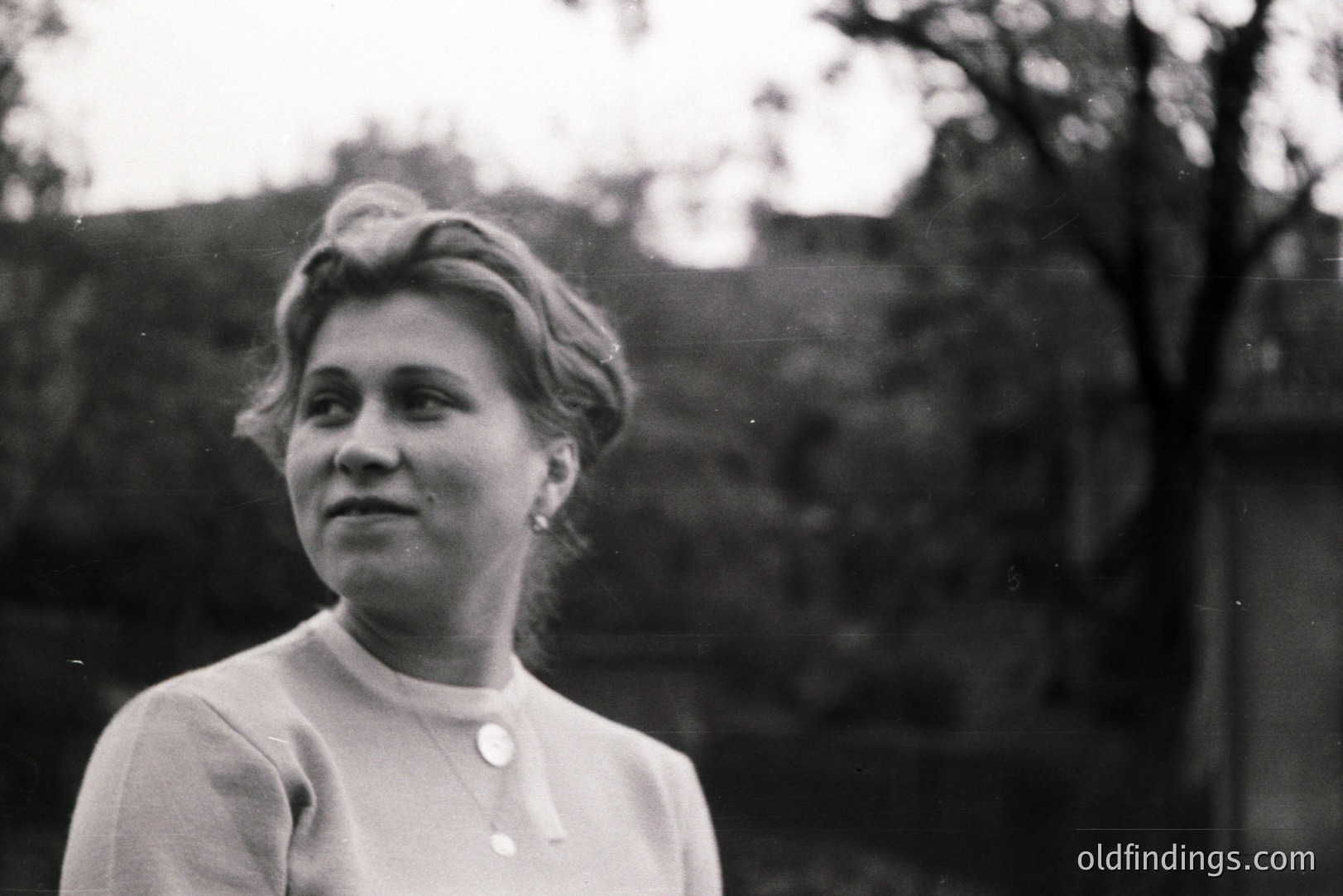 Portrait of a woman with a gently smiling expression, styled hair, and a simple, buttoned blouse. Captured outdoors, with blurred foliage as background. Likely a casual snapshot, suggestive of the mid-20th century (1950s-1960s). Offers potential for design or historical reference use.