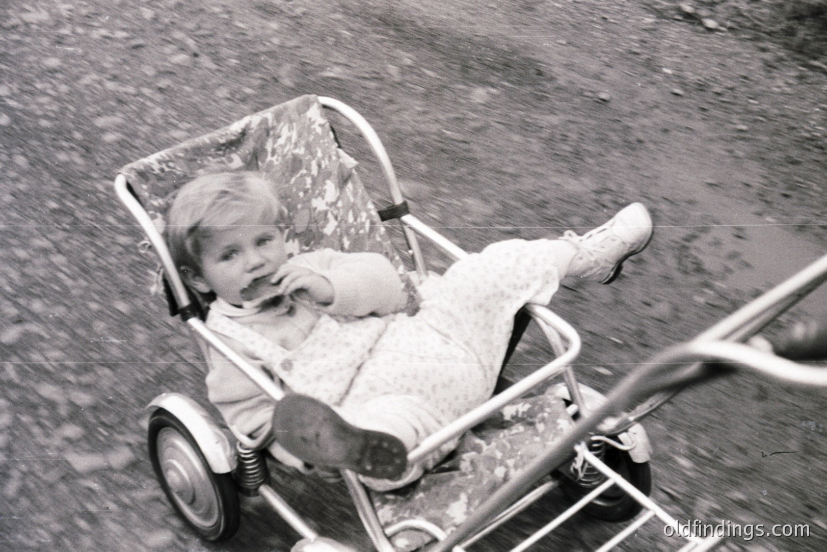A young child, likely a toddler, sits in a vintage chrome and fabric stroller. The child wears a patterned dress and boots, gazing slightly upwards, with food visible on their face. The stroller’s design suggests a mid-century aesthetic. A blurred figure is visible in the lower left.