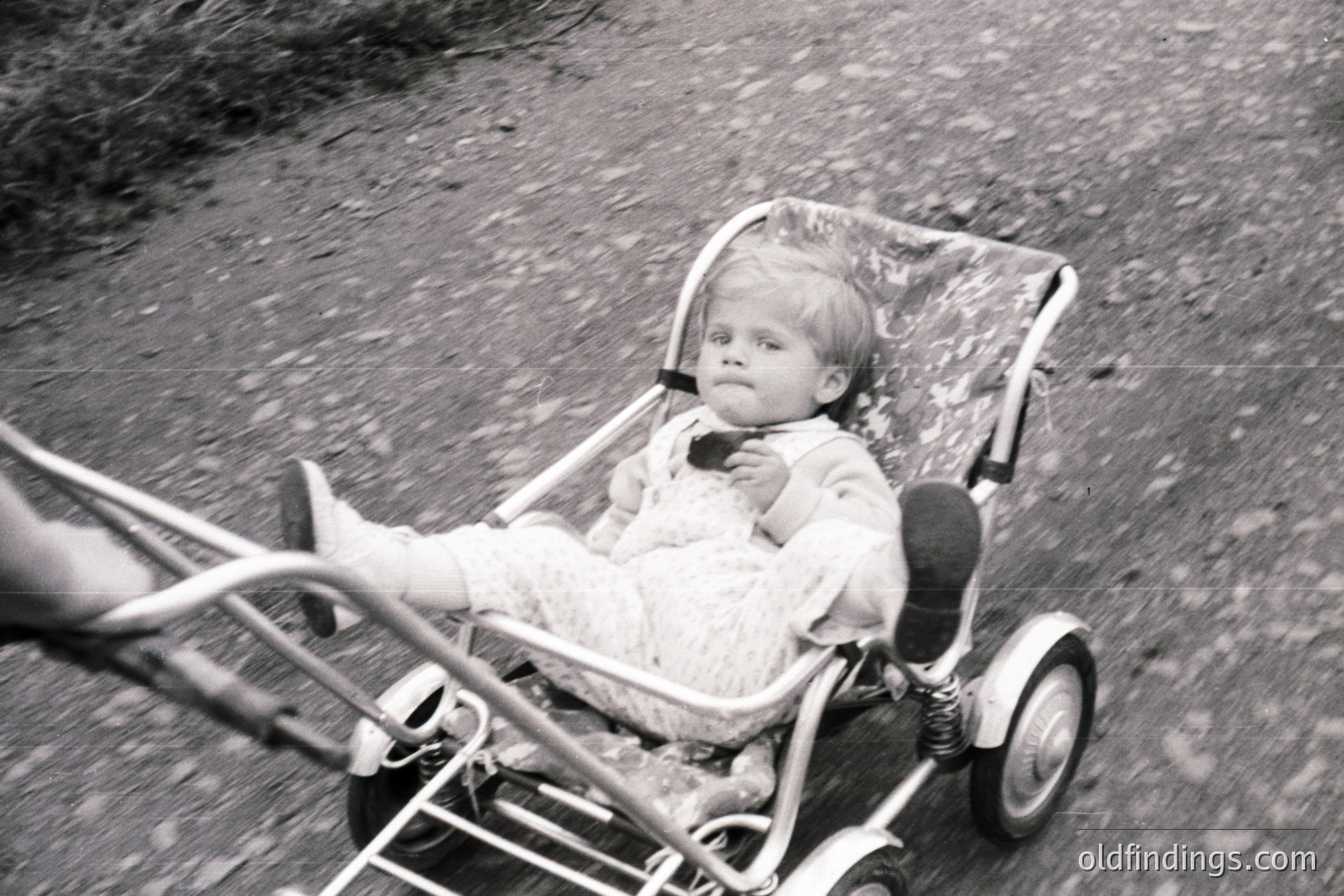 A young child sits in a vintage, metallic-framed wagon or stroller, captured in motion. The child wears a patterned knit outfit and appears to be looking downwards. The wagon's design suggests a 1960s or 70s aesthetic. Ground texture visible. Likely a snapshot, not a posed portrait.