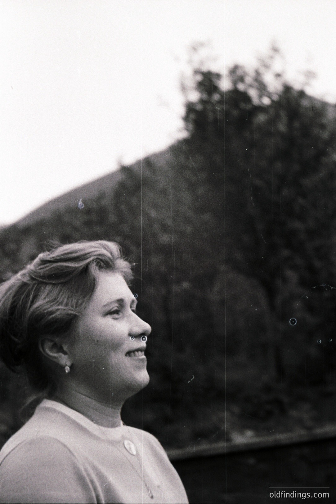 A young woman in profile smiles, gazing upwards. She wears a high-necked, textured sweater and simple earrings. The backdrop features a lush, hilly landscape with dense foliage. Likely mid-1960s style, possibly candid portrait. Valuable for fashion and design reference.