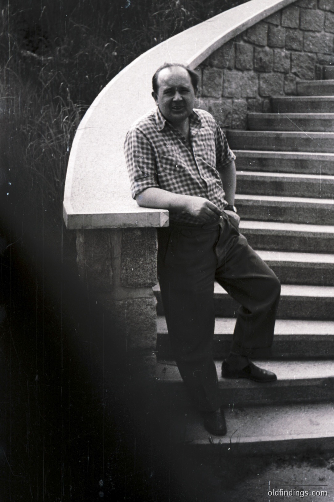 Man in a checkered shirt and slacks leans casually on a stone retaining wall beside a stone staircase. Appears to be a candid portrait, likely mid-1970s based on attire and photographic style. The stonework suggests possible European architecture. Could be stock for lifestyle or vintage design.